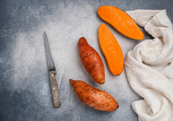 Raw organic whole and sliced sweet potatoes, a knife and a linen napkin on a gray concrete background. Fresh Root vegetables. Vegetarian and vegan food. Selective focus, top view and copy space