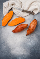Raw organic whole and sliced sweet potatoes, a knife and a linen napkin on a gray concrete background. Fresh Root vegetables. Vegetarian and vegan food. Selective focus, top view and copy space