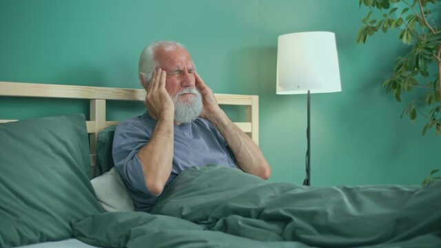 Tired Elderly Man Has Acute Headache While Sitting on ahe Bed At Home. Virus, Headache, Migraine, Anxiety. Seniors, Dizziness, High Blood Pressure, Health Care and Retirement Insurance.