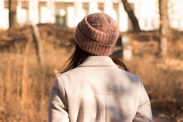 A girl with long dark hair in a coat and a knitted hat stands with her back to the camera against the background of an empty autumn park