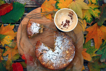 Top view on round oatmeal apple pie sprinkled with powdered sugar and a cardboard cup with cappuccino. Nearby lies a piece of cake from which a part has already been bitten off. - Powered by Adobe