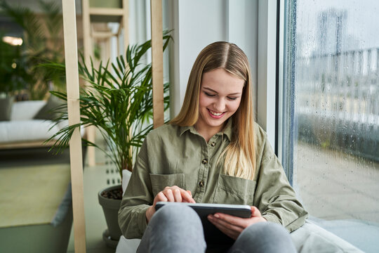 Smiling Female Intern Using Digital Tablet While Sitting By Glass Window In Office