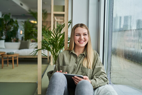 Smiling teenage female intern with digital tablet looking through window while sitting in office