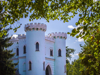 Fototapeta premium Part of a beautiful fairytale fortress surrounded by foliage and trees on a sunny day. The walls of the old building are painted in light blue and white. Magical atmosphere.