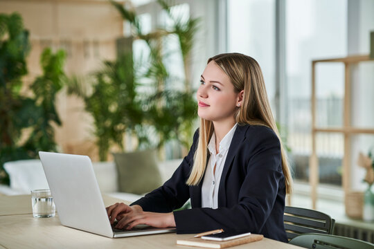 Thoughtful teenage intern looking away while sitting with laptop at desk in office
