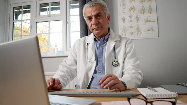 Portrait Of Mature Male GP Wearing White Coat At Desk In Doctors Office