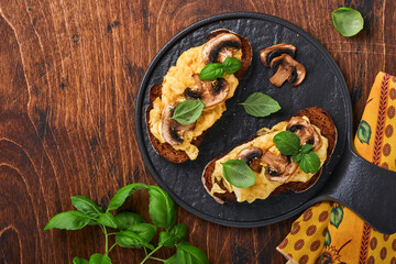 Scrambled eggs with fried mushrooms and basil on bread on black table background. Homemade breakfast or brunch meal - scrambled eggs and mushrooms sandwiches. Top view with copy space