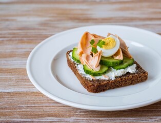 Smorrebrod, an open sandwich with boiled salmon, cream cheese, quail egg and cucumber slices on a white plate on a wooden background. Sandwich recipes.