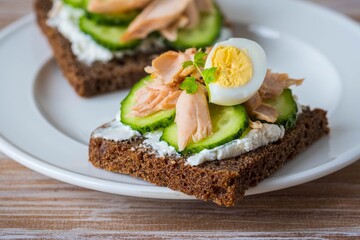 Smorrebrod, an open sandwich with boiled salmon, cream cheese, quail egg and cucumber slices on a white plate on a wooden background. Sandwich recipes.