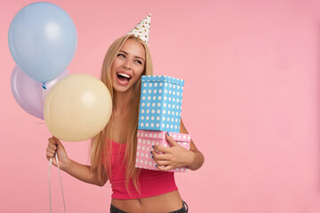 Studio shot of young blonde woman wears red top, posing against pink background, holding balloons and gifts. Isolated