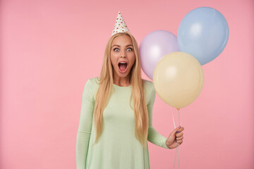 indoor portrait of young blonde female, wears green dress, party hat posing over pink background with shocked, excited facial expression. isolated