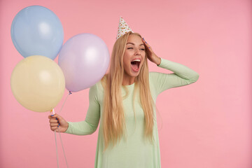 indoor portrait of young blonde female, wears green dress, party hat posing over pink background with shocked, excited facial expression. looks aside. isolated