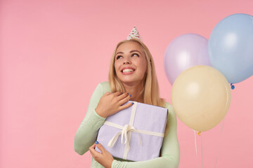 indoor portrait of young blonde female, wears green dress, party hat hug her big gift box and feels happy and excited posing over pink background