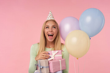 indoor portrait of young blonde female, wears green dress, party hat holding few gifts, looks upwards with happy facial expression. posing over pink background