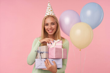 indoor portrait of young blonde female, wears green dress, party hat holding few gifts, smiles broadly and feels satisfied, posing over pink background
