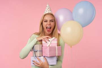 indoor portrait of young blonde female, wears green dress, party hat holding few gifts, smiles broadly and feels satisfied, posing over pink background
