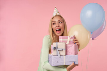 indoor portrait of young blonde female, wears green dress, party hat holding few gifts, smiles broadly and feels satisfied, looks aside posing over pink background