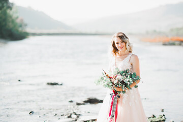 Fototapeta na wymiar Beautiful brunette bride in elegant white dress holding bouquet posing near river