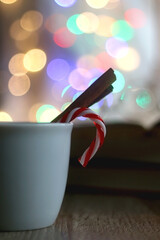 Cup of warm drink with cinnamon stick and candy cane, open book, reading glasses, lit candles and colorful bokeh lights. Selective focus.