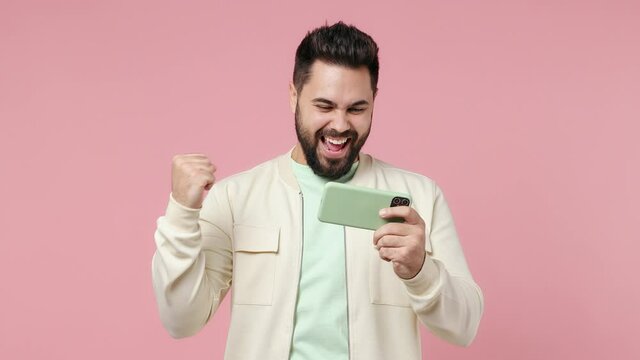 Gambling young bearded brunet man 20s wears white shirt using play racing on mobile cell phone hold gadget smartphone for pc video games isolated on plain pastel light pink background studio portrait