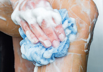 Young woman washing legs in shower bath with blue sponge and soap foam