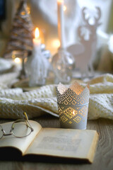 Fototapeta na wymiar Open book and reading glasses on the table. Lit candles, soft blanket and Christmas decorations in the background. Hygge at home. Selective focus.