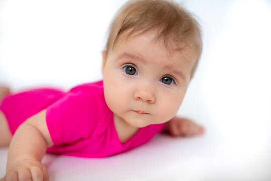 cute, beautiful little girl in a pink bodysuit
 and a bandage lies on a white background, smiles and gnaws toys. beautiful baby. teeth are erupting. portrait of a baby. portrait of a beautiful girl