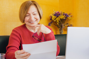 Happy middle aged elderly woman reading paper document or postal letter from friend, sitting at home indoor. Reaction of pleasant paperwork bank notice or post correspondence.Taking bank loan concept