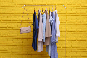 Rack with different stylish clothes and bag near yellow brick wall