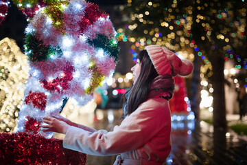 A young Korean Asian woman in a beige coat and hat on a walk through the winter night Christmas city, decorated with neon lights, garlands and lots of colorful light bulbs