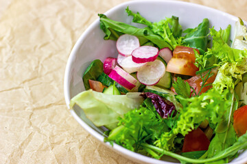 Bright fresh green salad leaves with red tomatoes, cucmbers and radish in the white bowl on light background.