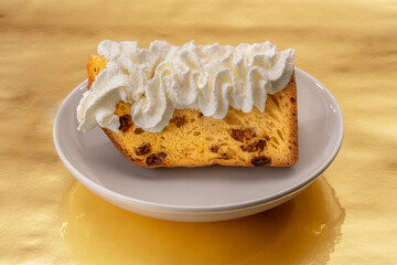 Slice of panettone cake with whipped cream in white plate on a golden background, typical Italian Christmas dessert