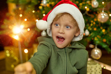 Cute boy in a New Year is hat, with big blue eyes and a mouth open in surprise holds a sparkling...