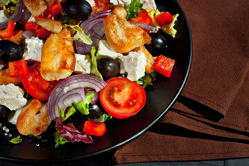 Salad with tomatoes, peppers, olives and chicken fillet. Fresh salad in a black plate on a dark background. Brown towel near the plate.