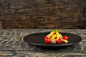 Pasta with meat and tomatoes in a black plate. Pasta on an old wooden table. Dish on a shabby brown background. Copy space and free space for text near food.