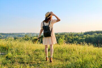 Back view, woman with backpack in hat enjoying summer landscape of wild nature