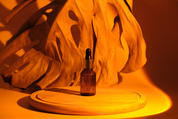 Glass vial bottle of essential oil or serum for facial care on a wooden round podium in the light of a sunset lamp. In the background, a dried brown monstera leaf