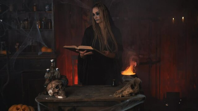 Halloween, witch reading magic book use cauldron prepare poison or love potion. Black magic occult female wizard in dark room cage spider web human skull Necromancer. voodoo. Handheld Blow kiss