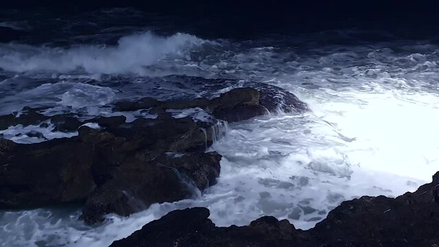Sea waves splash and crash into rocks with much white foam. Night cinematic shot sea violently splashing over the rocky coast, highlighted by strong light, night video