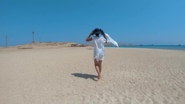 Fit woman ini white dress and hat walk on paradise beach at Giftun island in Hurghada, Egypt with relaxing scarf waving on breeze, Slow motion