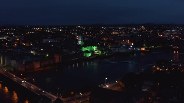 Fly above night city, green illuminated large building on Shannon river waterfront. Limerick, Ireland