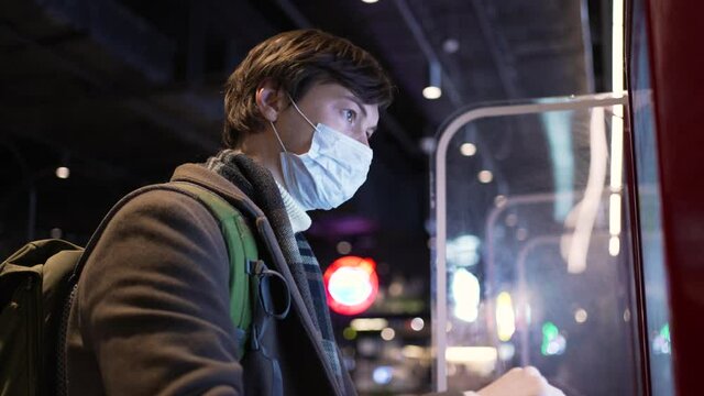 Young brunet man wearing medical mask and with backpack orders travel pass via ticket vending machine standing at bus stop in evening