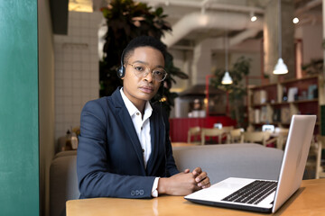 Serious African American woman uses a headset for a video call, works in a cafe, during a tourist business trip, business woman focused and thoughtful at work