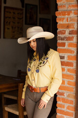 A teenager in cowgirl clothes leaning against a brick wall in a ranch