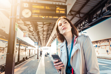 Young woman with departure times behind her waiting for her train while holding her mobile phone -...