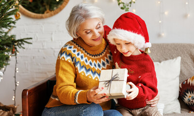 Fototapeta na wymiar Little boy grandson giving Christmas gift box to smiling grandmother during winter holidays