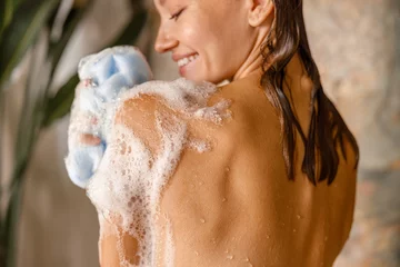 Fototapete Spa Happy young woman applying shower gel on her body using loofah sponge while taking shower. Spa and body care concept