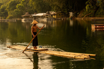 a fisherman in the countryside is crossing a lake on a bamboo raft - obrazy, fototapety, plakaty