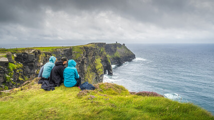 Group of young people sitting on the edge of cliff on iconic Cliffs of Moher, popular tourist...