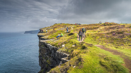 Group of people or tourists hiking and sightseeing iconic Cliffs of Moher, popular tourist attraction, Wild Atlantic Way, County Clare, Ireland - obrazy, fototapety, plakaty